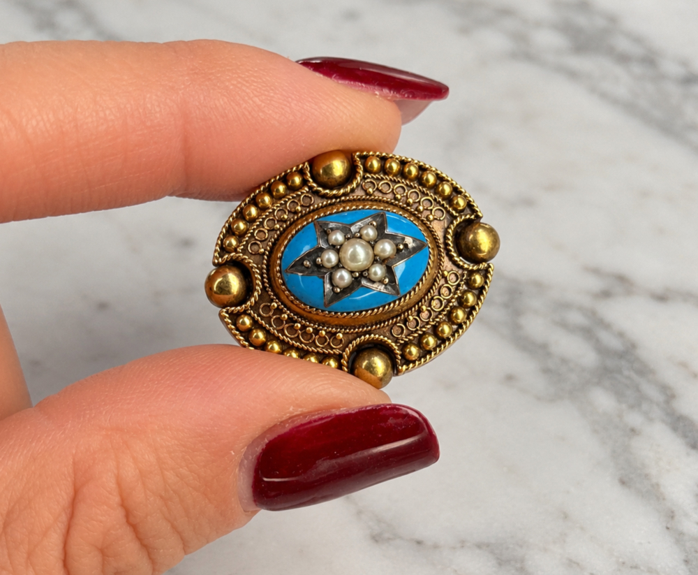Two fingers holding an antique Victorian Etruscan Revival gold and enamel brooch with seed pearls in a star setting